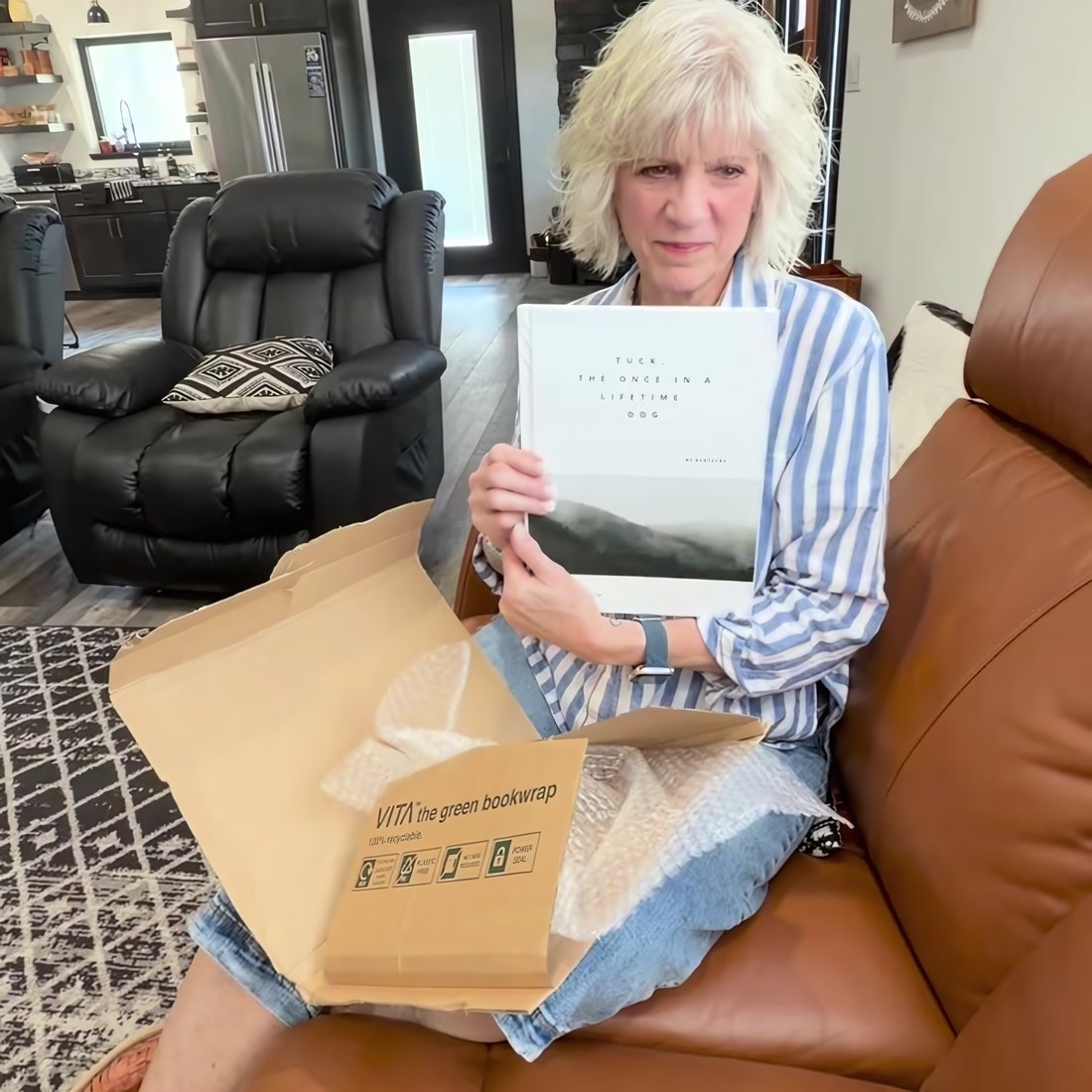 A woman on her sofa holding up her Rowen Hollow book, the shipping box open on her lap